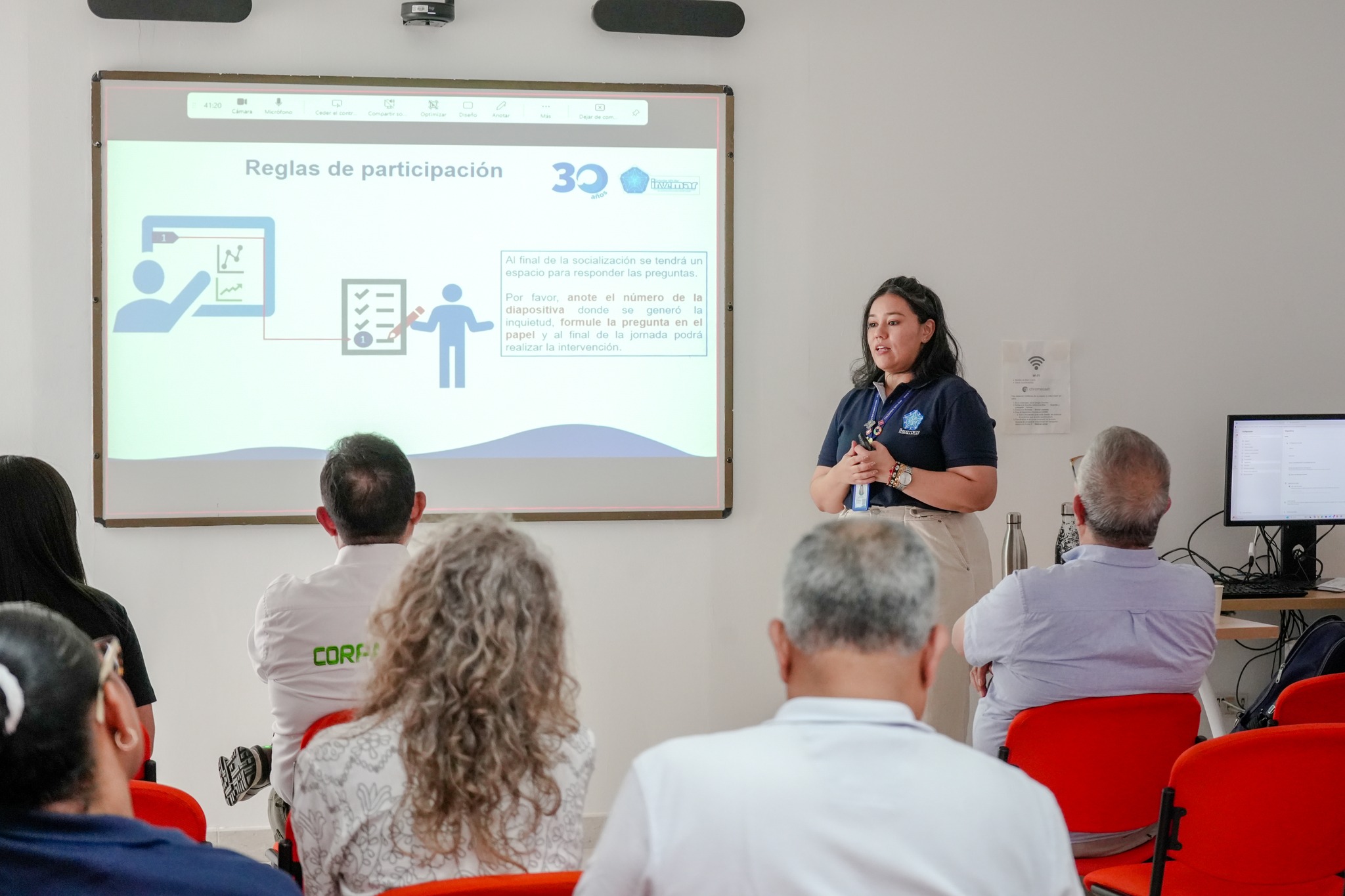 Persona de pie dando una presentación frente a un grupo de asistentes sentados, con una diapositiva proyectada en una pantalla que muestra reglas de participación.